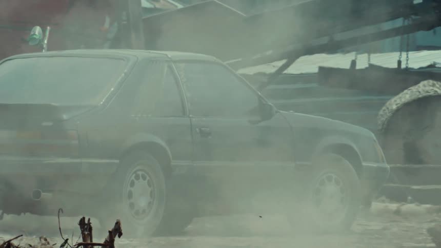 A vintage sports car is parked in a dusty and smoky junkyard as the camera slowly zooms out