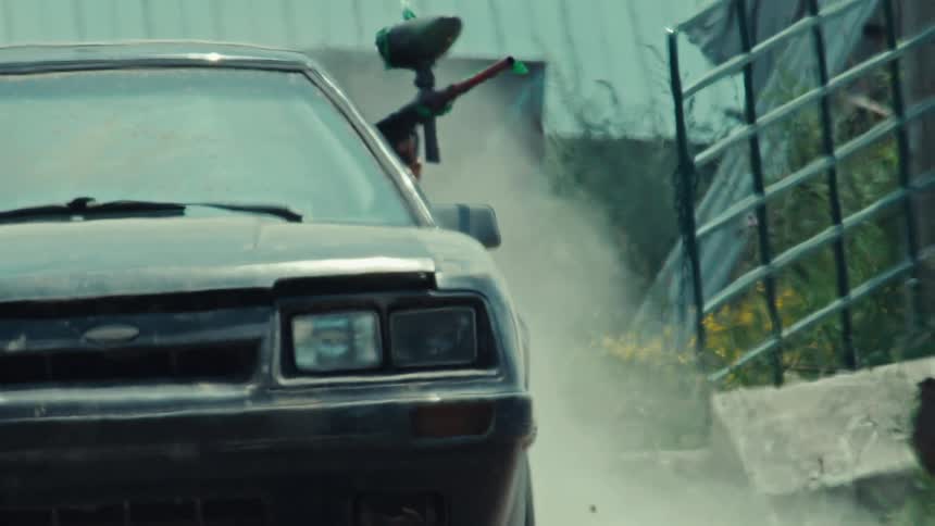 A paintball player leans out of a classic Ford Mustang with a marker aimed as the car kicks up dust in slow motion