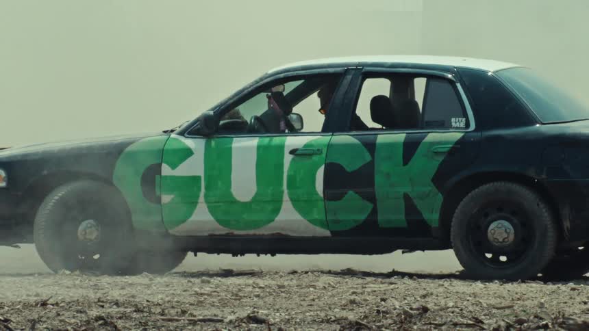 Side view of a stationary black sedan with rebellious green graffiti kicking up dust in a desolate, arid landscape