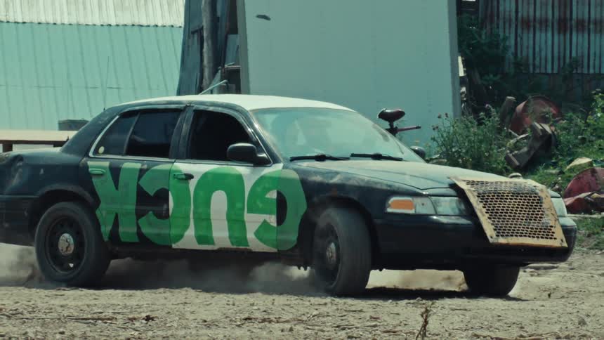 A demolition derby car with 'GUCK' spray-painted on the side does a burnout in slow motion in a dusty junkyard