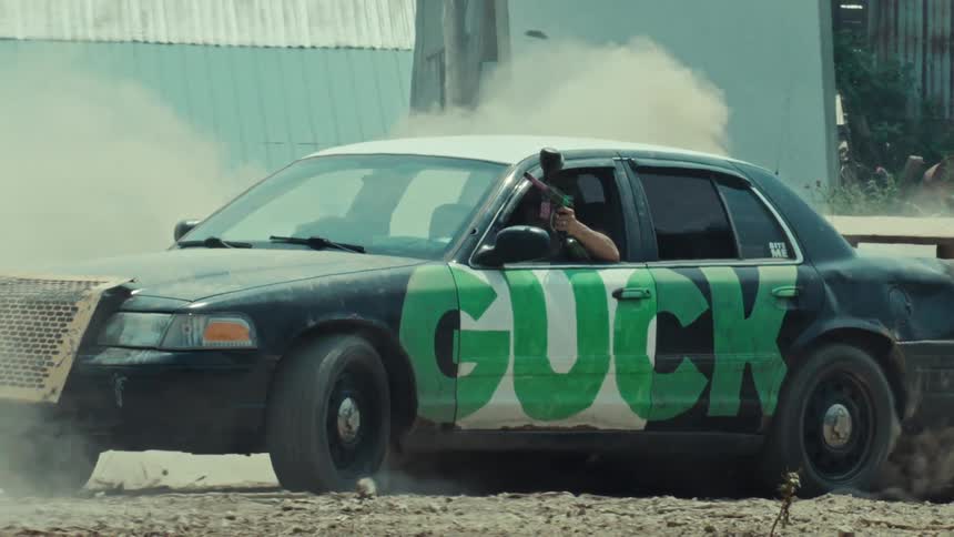 A man in a modified car does a burnout while aiming a paintball gun out the window in slow motion