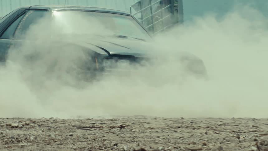 A dark sports car is shrouded in thick smoke as it performs an intense burnout in dramatic slow motion