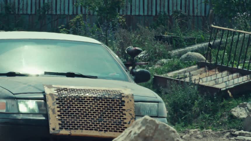 A paintball player in camouflage takes cover behind a fortified police cruiser in an abandoned junkyard during a match