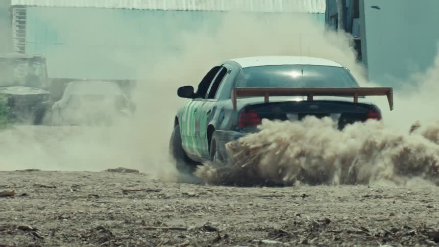 A customized black and white car with a spoiler does a burnout on a dirt track, kicking up a large cloud of dust in slow motion