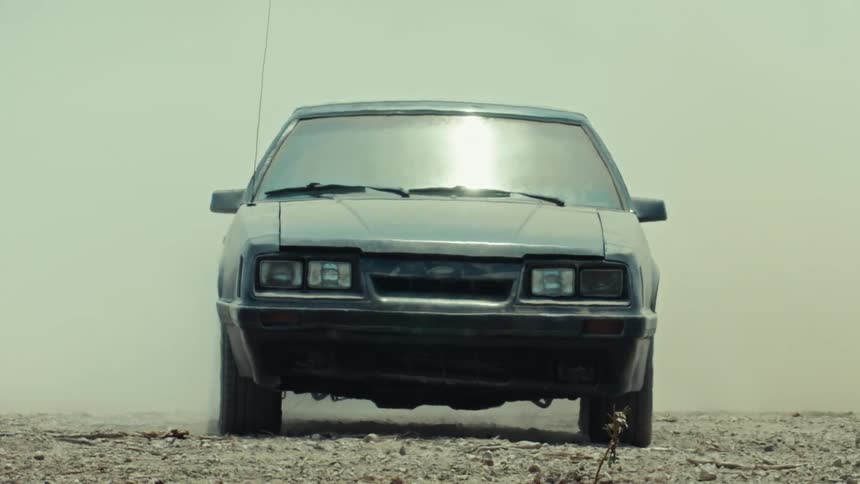 A vintage black car sits idle on a dusty road in a hazy, hot, and desolate environment