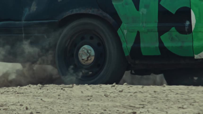 A slow-motion close-up shows a demolition derby car's flat tire spinning and kicking up a large cloud of dust on a dirt track