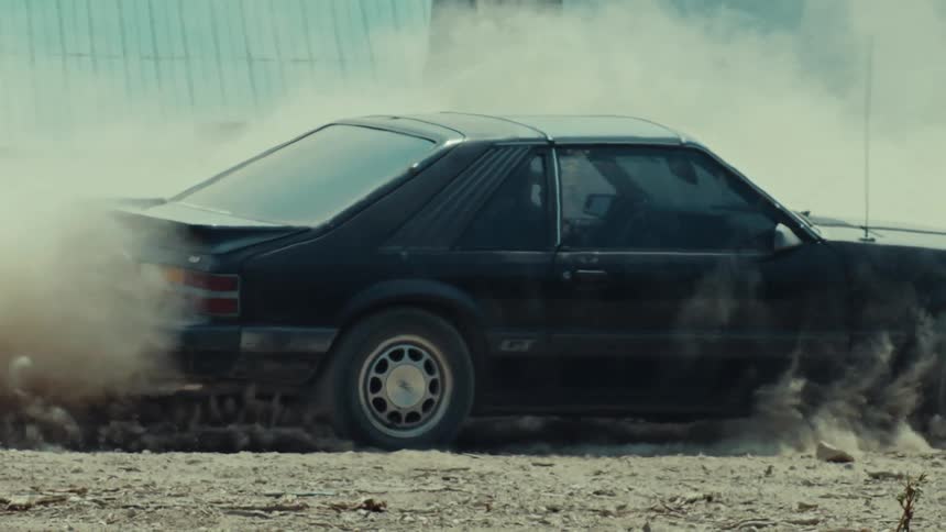 Side view of a vintage black muscle car doing a burnout on a dirt road, kicking up a huge dust cloud in slow motion