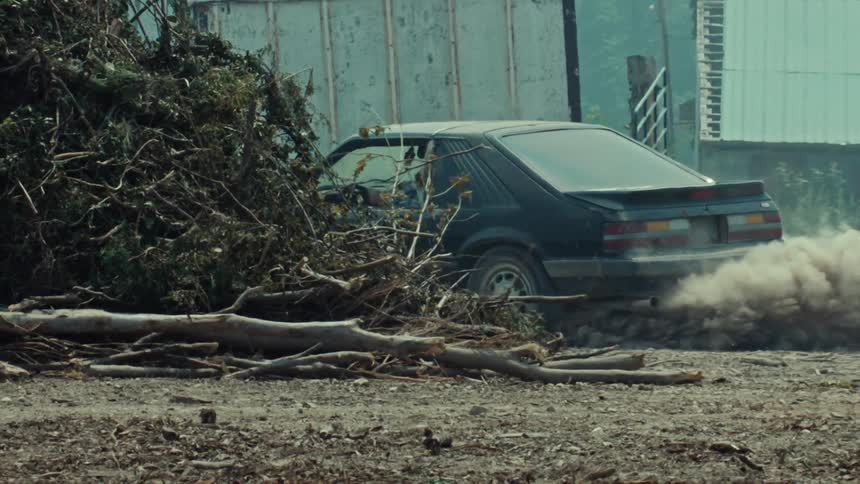 A man in a classic black muscle car performs a powerful burnout in slow motion, disappearing into an explosive cloud of dust and dirt
