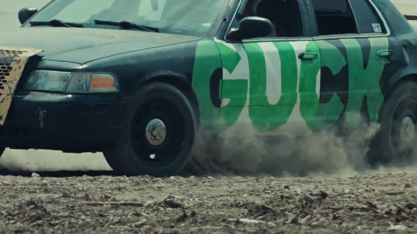 A modified black sedan with green graffiti does a burnout in slow motion, its wheels spinning and kicking up a large cloud of dust from the dirt road