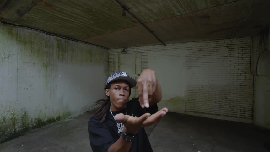 A young man with dreadlocks performs intricate finger tutting dance moves in a gritty, empty concrete room
