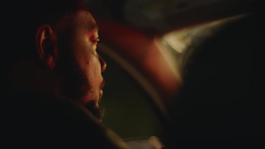 A serious man sits behind the wheel of his car at night, his face illuminated by the passing red and white city lights