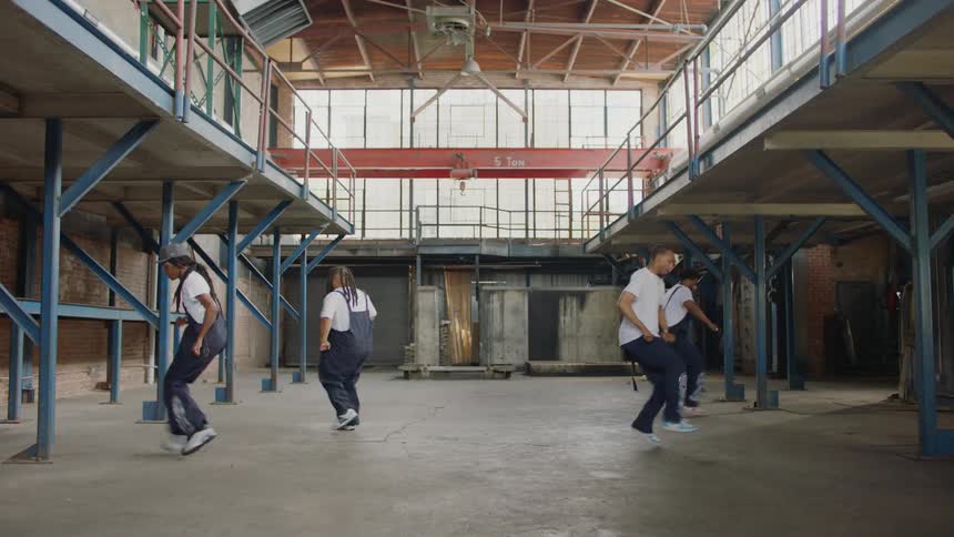 A diverse group of four talented dancers in matching overalls perform a synchronized street dance routine in a large, empty industrial warehouse