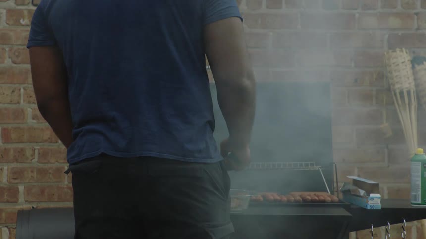 A man tends to sizzling sausages on a smoky barbecue grill against a brick wall, filmed in slow motion