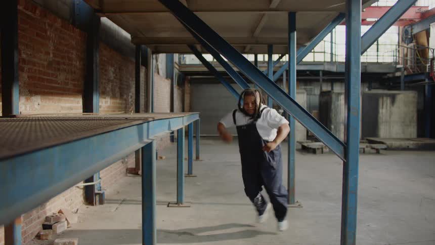 Listening to music on headphones, a man in overalls breaks into an expressive and energetic dance in an industrial warehouse