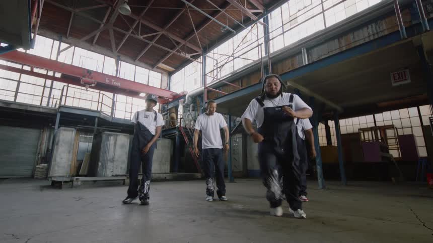 A diverse dance crew performs an energetic hip-hop routine in a large, empty industrial warehouse
