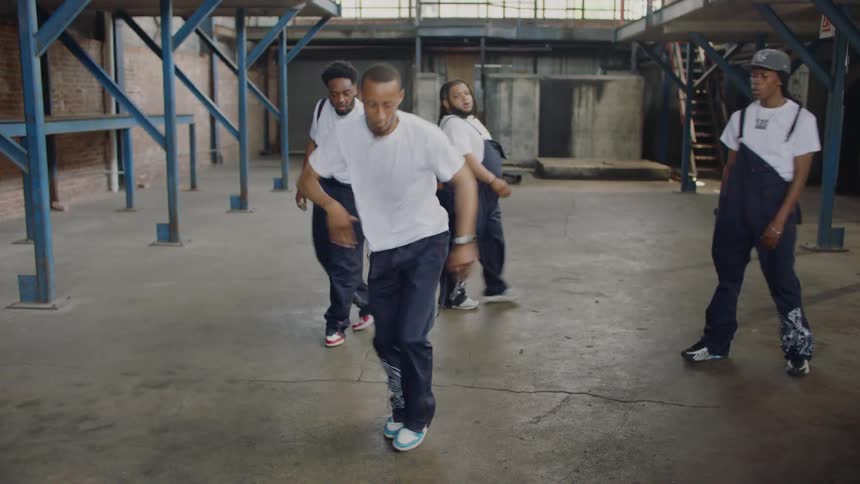A talented group of diverse dancers in matching overalls perform intricate hip-hop choreography in an urban warehouse