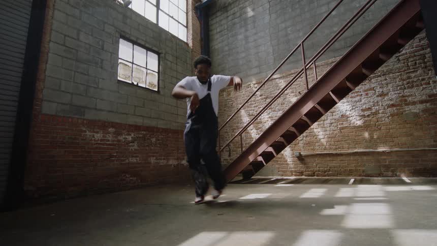 A talented man in overalls performs an energetic and expressive hip-hop dance routine inside a large, empty industrial warehouse
