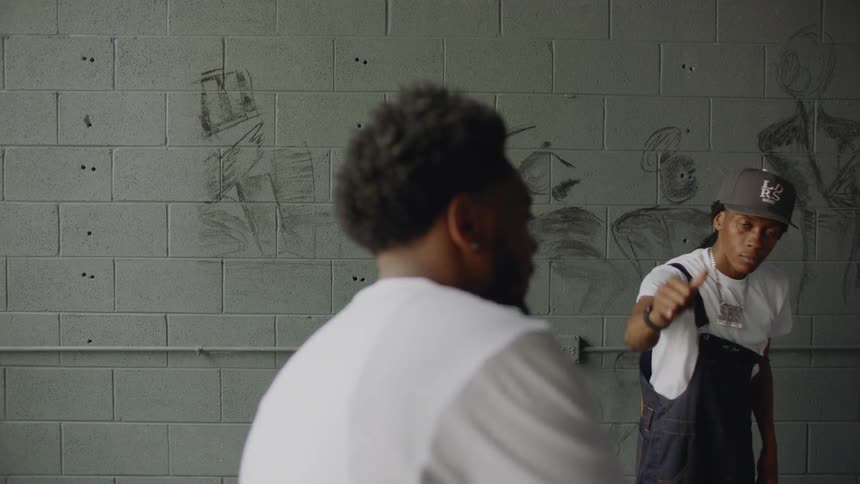 In a slow-motion shot with a rack focus, a young man with a cigar smiles and shows off his grillz while his friend dances behind him against a graffiti wall