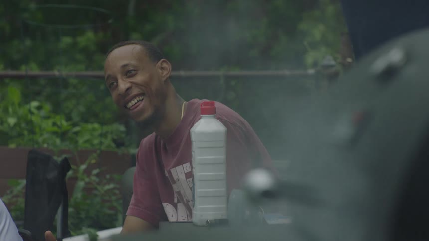 A happy young man smiles and socializes with friends at a backyard barbecue as smoke gently rises from the grill