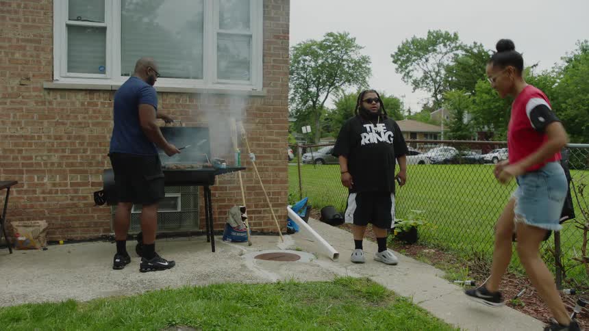 A man grills on a smoker while his friends dance and enjoy themselves at a backyard barbecue