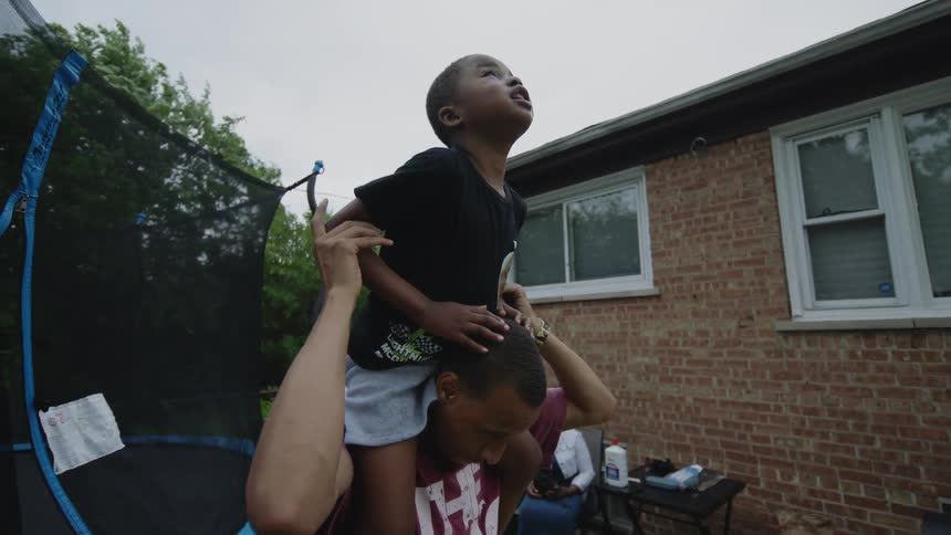 A loving father playfully lifts his young son off his shoulders and holds him upside down while smiling at a backyard family party in slow motion