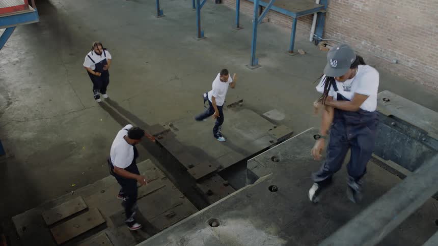 A high-angle view shows a group of four street dancers performing a powerful, synchronized routine in an empty factory