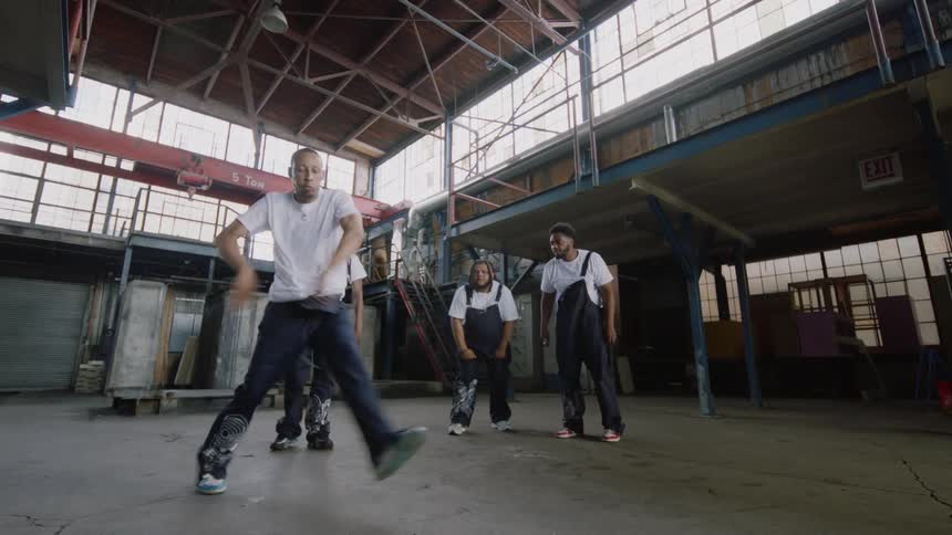A talented dancer performs an energetic solo routine for his crew inside a large, empty industrial warehouse
