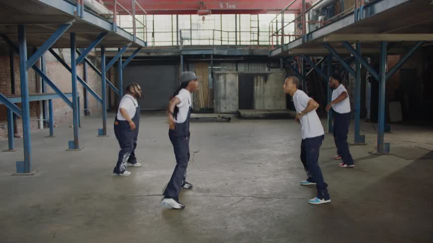 A crew of dynamic dancers perform a synchronized hip-hop routine inside a gritty industrial warehouse