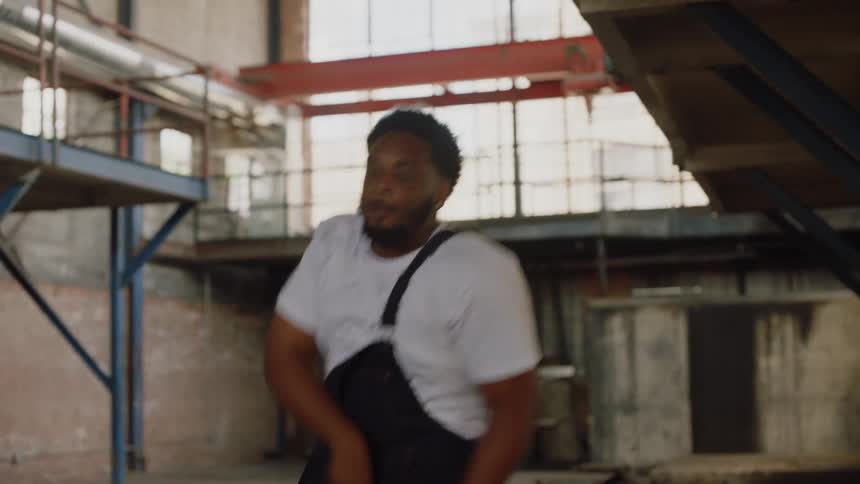 A skilled young man in overalls performs an energetic and expressive dance routine in a large industrial warehouse