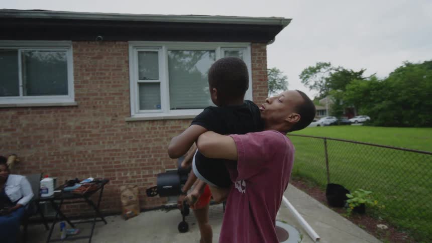 A loving father playfully holds and kisses his young son in his arms during a family backyard cookout in slow motion