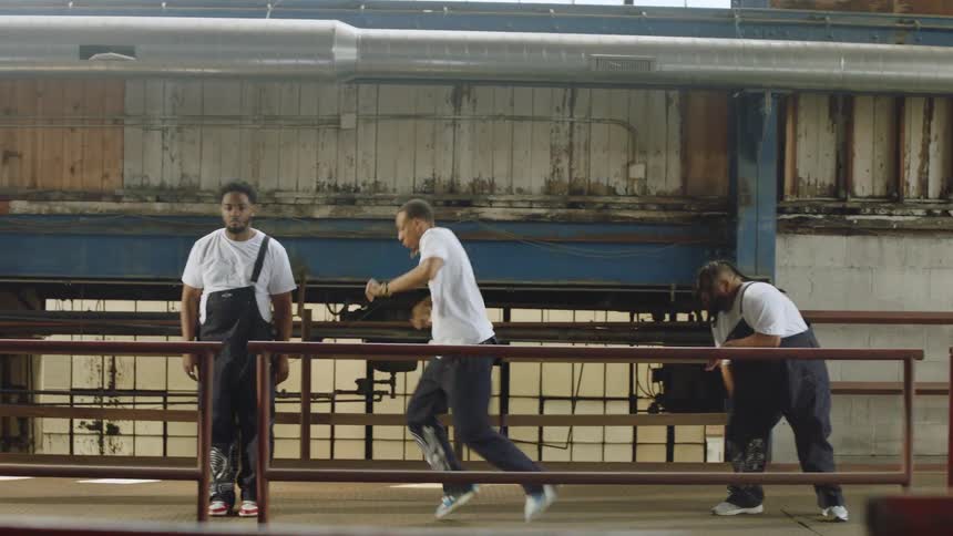 A diverse group of male dancers in overalls energetically performs a hip-hop routine on a metal walkway inside a large, gritty industrial warehouse