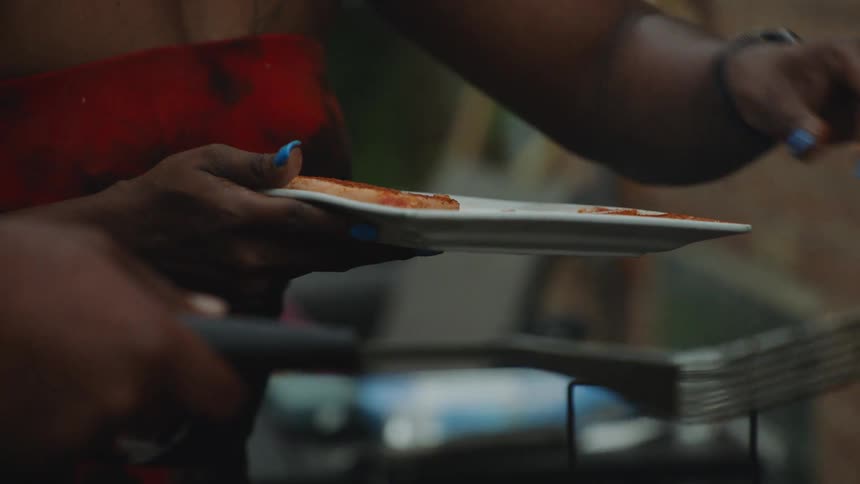 A slow-motion close-up of a woman preparing a plate of food at a backyard barbecue with meat sizzling on the grill