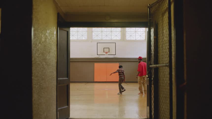 A father coaches his young son as he practices dancing in an empty school gymnasium, viewed from a dark hallway