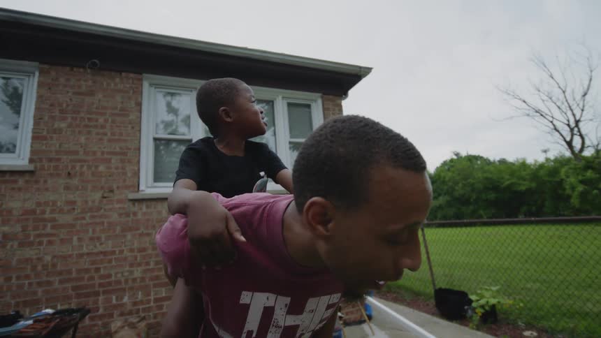 A loving Black father playfully lifts his young son onto his back for a piggyback ride during a family cookout in their backyard