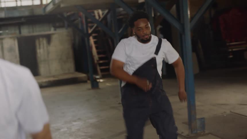 An energetic Black man in overalls performs a powerful and expressive dance routine in a raw, industrial warehouse space