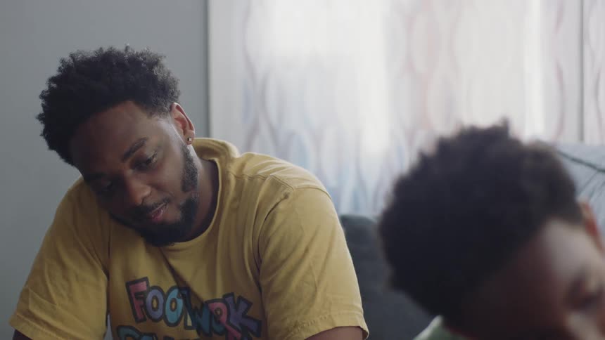 A young father's joyful smile slowly fades to a look of concern as he watches his son in their living room