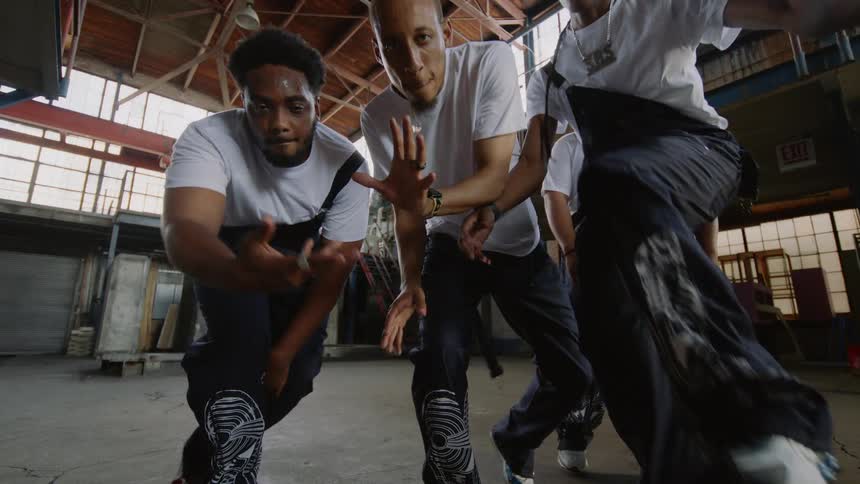 A crew of male dancers performs an energetic synchronized hip-hop routine in slow motion inside a spacious industrial warehouse