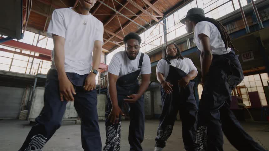 A group of male dancers perform a synchronized routine in slow motion inside a large industrial warehouse