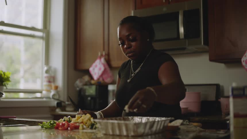 A woman carefully seasons and mixes freshly chopped vegetables in a pan in her sunlit kitchen