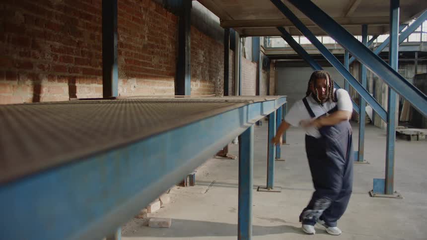 An energetic man with dreadlocks wearing headphones and overalls dances freely in a large, empty industrial space