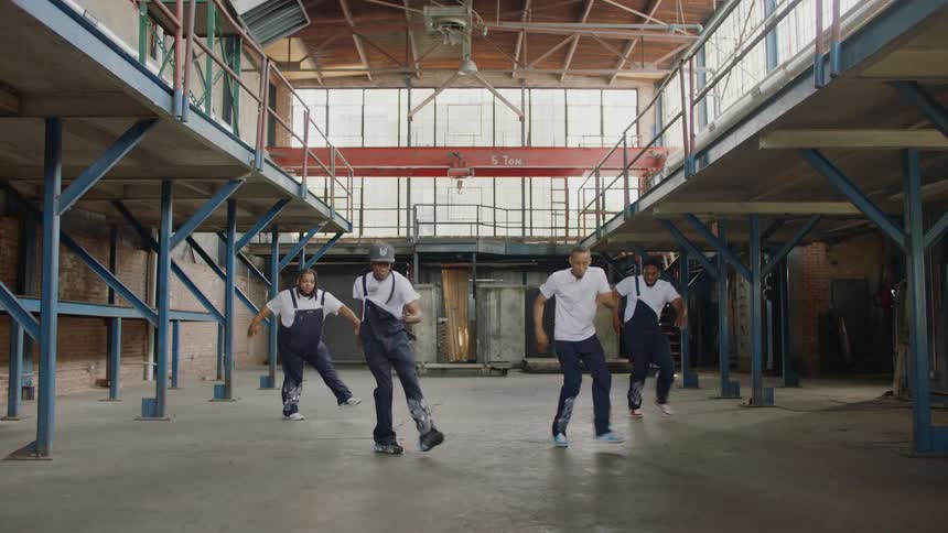 A crew of four dancers performs a synchronized routine in a large industrial warehouse