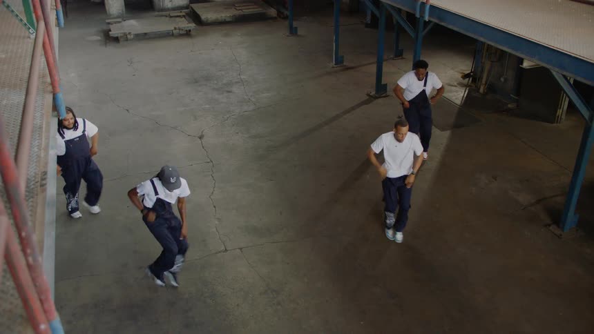 From a high angle, a talented group of four street dancers in matching overalls performs a synchronized routine inside a spacious industrial building