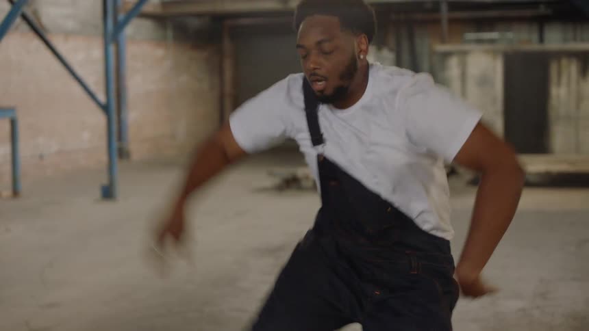 A talented Black male dancer performs an intense and energetic street dance routine inside a large, empty industrial warehouse