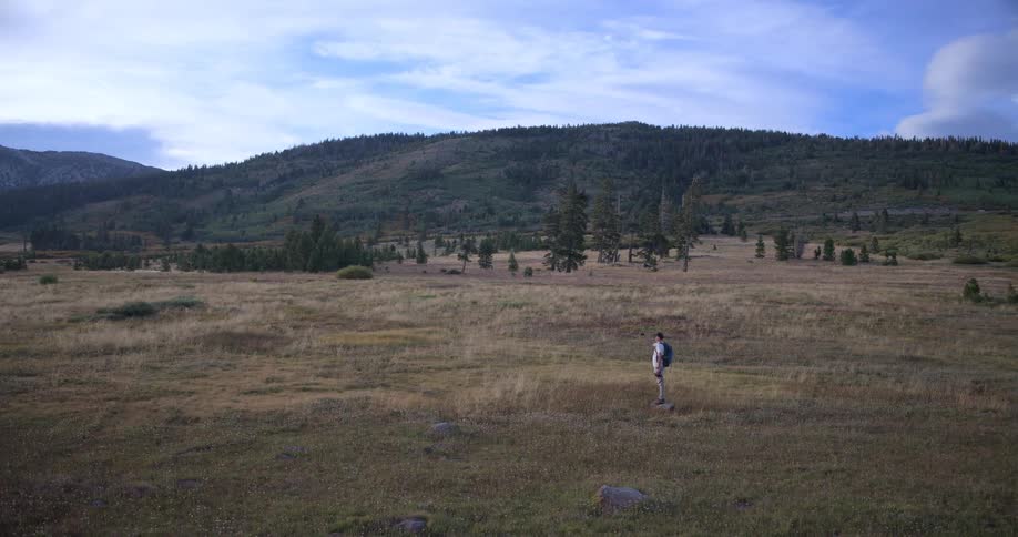 A lone hiker with a backpack walks into a vast mountain meadow and pauses to admire the scenic landscape