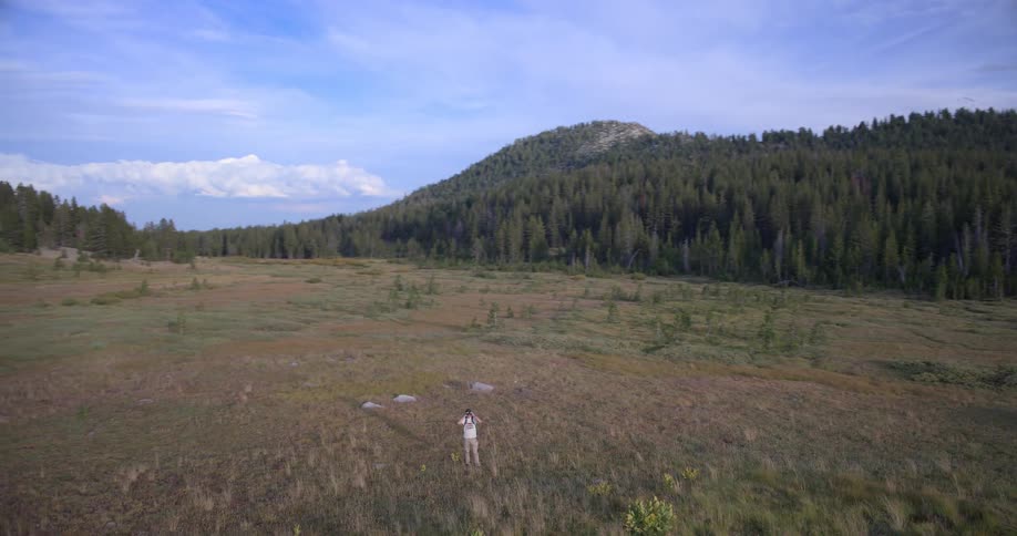 An adventurous hiker with a backpack explores a vast mountain meadow as the camera slowly rises to reveal the breathtaking and immense landscape