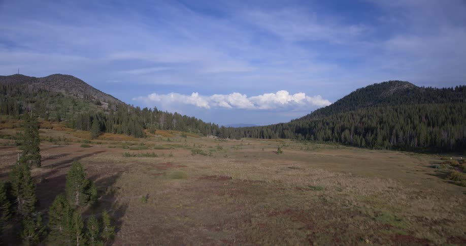 An aerial view slowly drifts over a sprawling mountain meadow under a partly cloudy sky
