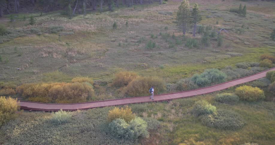 An aerial view shows a lone hiker with a backpack walking along a wooden boardwalk through a scenic mountain meadow at sunrise