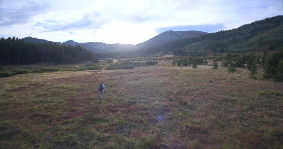 Slow motion panning shot of a man with a backpack hiking through a vast, scenic mountain meadow surrounded by forests and hills