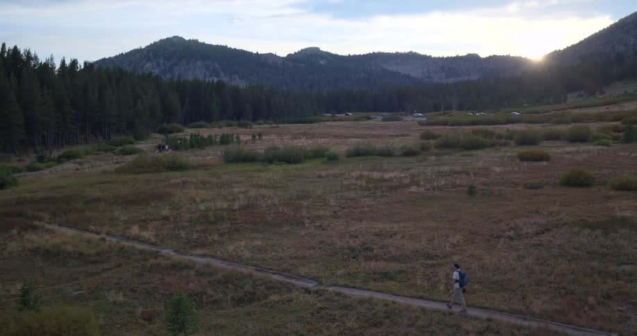 Aerial view of a lone hiker with a backpack walking along a trail in a vast mountain meadow at sunset