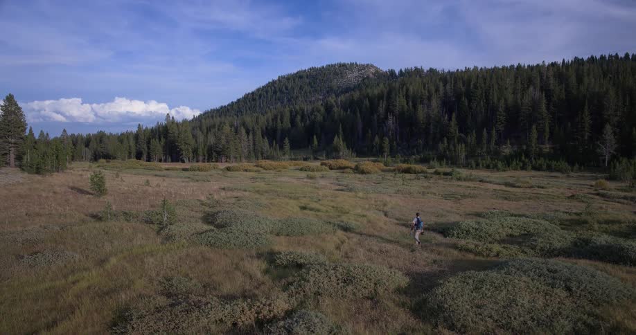 A lone hiker with a backpack and trekking pole walks in slow motion across a vast mountain meadow under a blue sky
